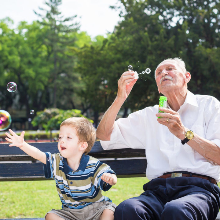Grandfather blowing bubbles on park bench with young toddler boy